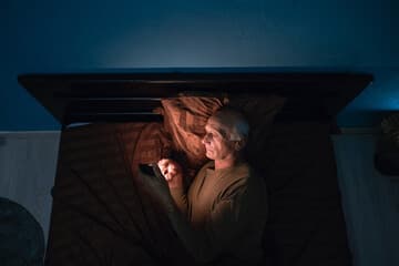 A smartphone glowing in a dark room with a blurred scroll feed and a silhouetted figure.