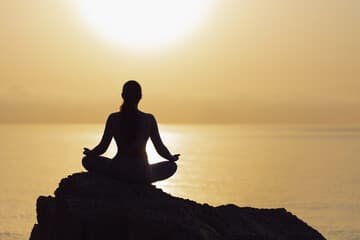 Silhouette of a person meditating in lotus position on a rock against a golden sunset over the ocean