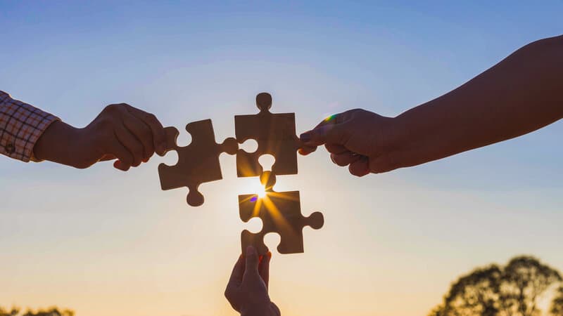 Three pairs of hands fitting wooden puzzle pieces together against a sunset sky, symbolizing cooperative problem-solving over zero-sum competition