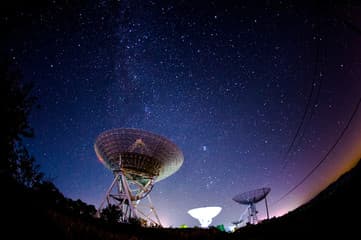 A satellite dish array at night with three faint colored light beams converging.