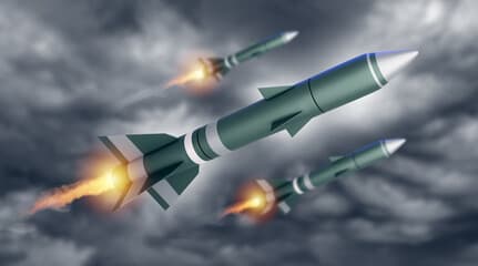 A naval vessel being loaded with missile systems at a European shipyard under overcast sky.