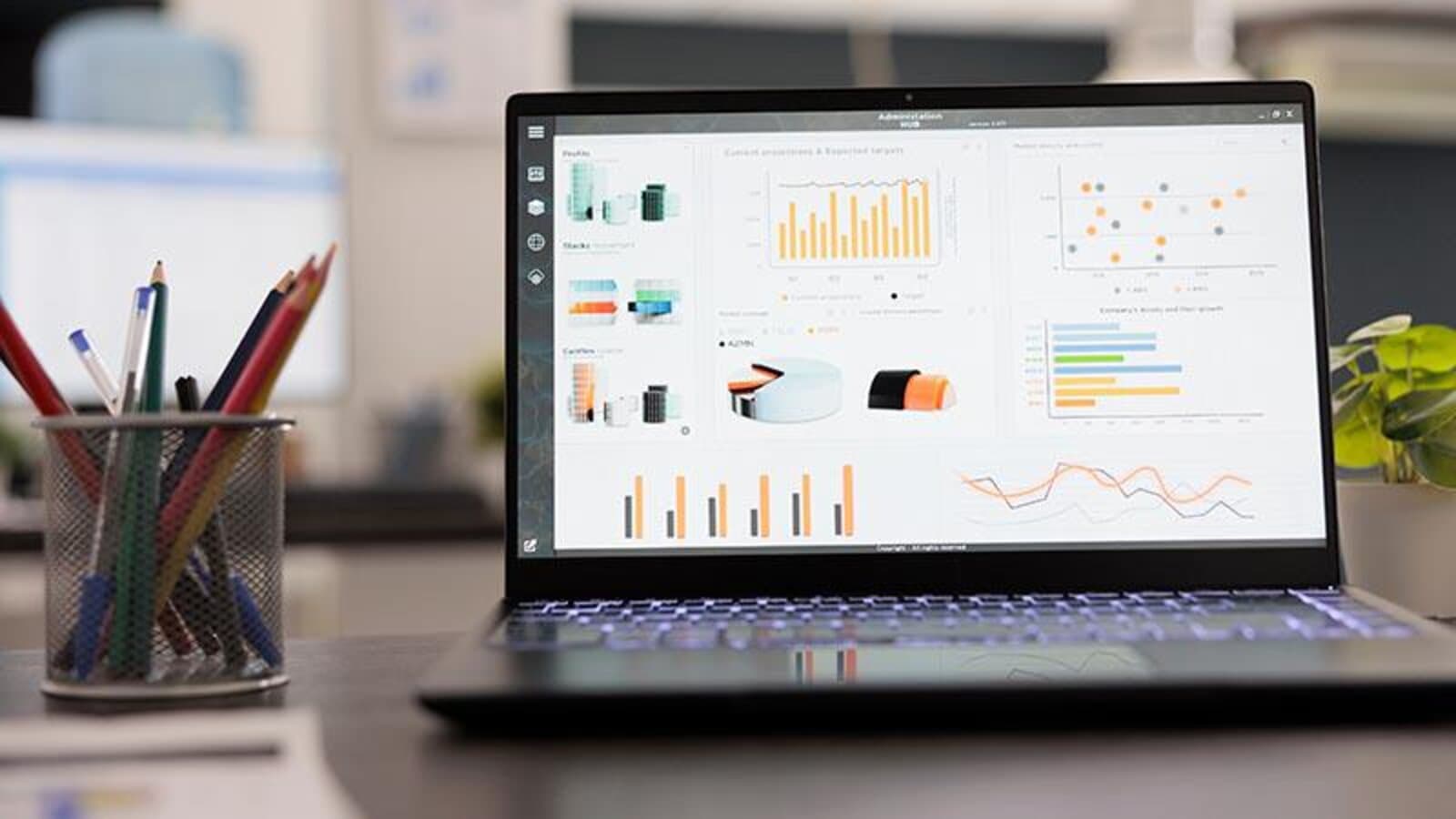 A laptop on a desk displaying financial charts and cost analysis graphs, with a cup of pencils nearby