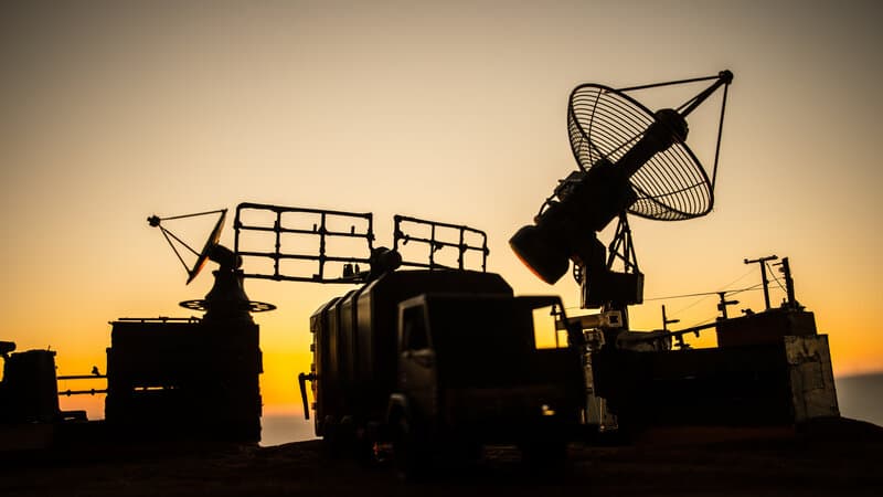 A desert military outpost at night with searchlight beams crossing the sky and a distant glow on the horizon
